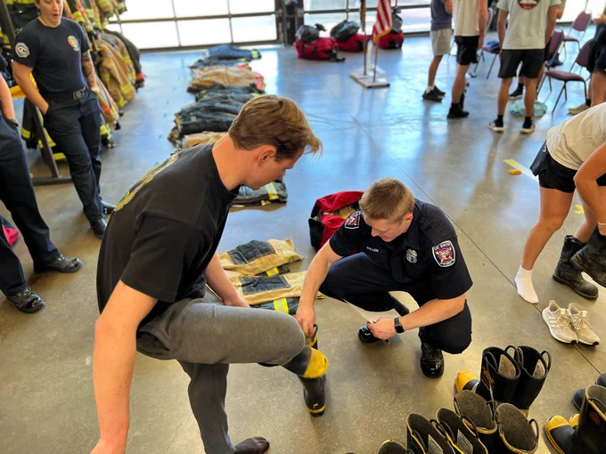 SFF K. Cullen in navy blue UCDFD uniform assisting a 2024 SFF Recruit wearing a black shirt and grey sweatpants try on wildland boots.