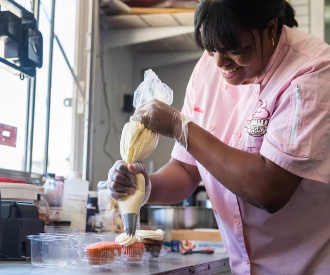 A woman wearing a pink chef's coat frosts cupcakes. 