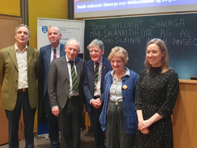 Cúigear ball d'Acadamh na Lianna agus Oifigeach Gaeilge TCD ag seasamh os comhair comhartha agus clár dubh 