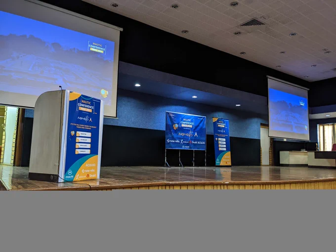 A conference room stage with two presentation screens, Mautic Conference India 2024 promotional banners, and podium.