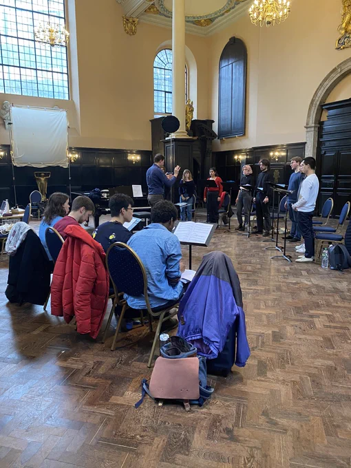8 singers rehearse in a spacious room with big windows and wood panelling while 4 Young composers look on from their seats