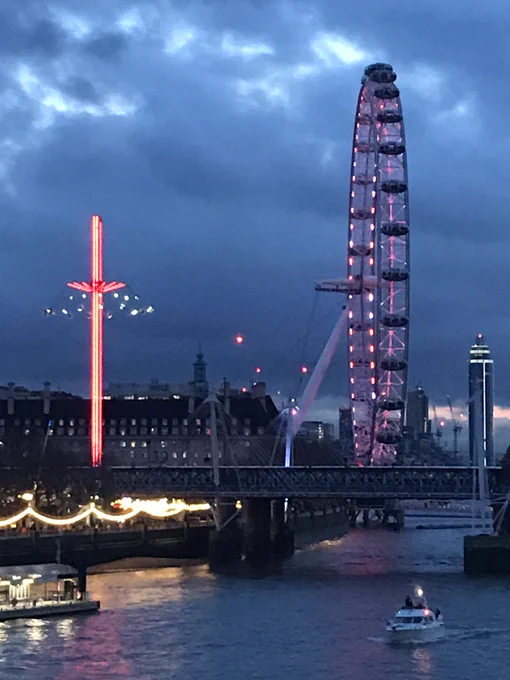 Photo of the river Thames at night. The sky is very dark blue and cloudy, with patches of light coming through the clouds. The London Eye (side angle view) is lit up pink/purple. The Southbank of the river (on left side) is lit with gold hanging lights along the bank, which is reflected on the river.  There's a motor boat coming towards the camera (bottom right). To the left of the photo and left of the London Eye, there's a fun fair ride which is a very tall vertical pole with chairs swinging around near the top of the pole and this is lit up in fluorescent pink and gold.