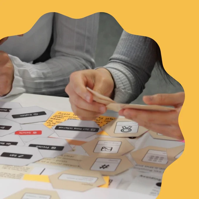 A photograph which takes up most of the image is placed towards the bottom left corner of the image. It depicts the hands of workshop participants looking through a selection of flashcards. On the table beneath the two pairs of hands in shot, there are more hexagonal cards laid out in a messy honeycomb-like shape. The image is cropped with curved, smooth, scalloped edges and is places on a bright mustard yellow background.