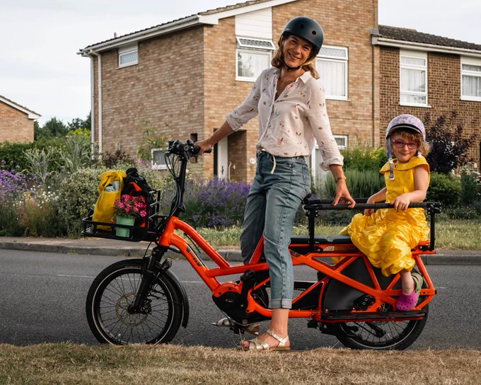 a blonde white woman wearing a black helmet, flowery blouse and blue jeans sat on the saddle of an orange electric cargo cycle with bags on the front and a child wearing a yellow dress, glasses and patterned helmet sat on the back. Both are located in the road in front of houses with plants in the background. 