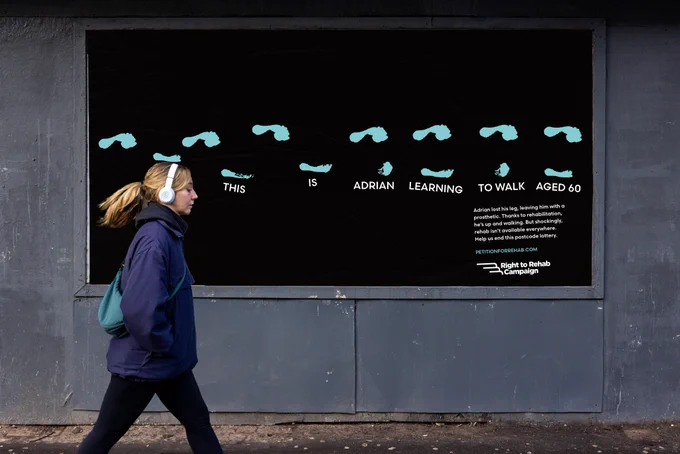 A woman walking past a billboard of the Right To Rehab campaign - the billboard is black and shows in bright coloured paint a patient's first footsteps during rehabilitation care with the text 'This is Adrian learning to walk aged 60'.
