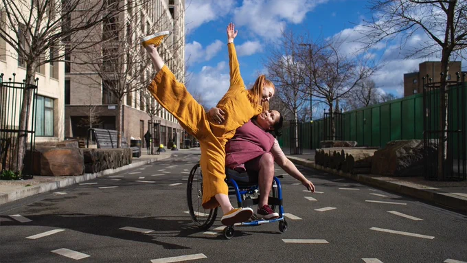 A wheelchair user in a brown dress dances with a non-wheelchair user in a yellow jumpsuit as they smile at each other in a supported dance-pose on a sunny street under a blue sky. 