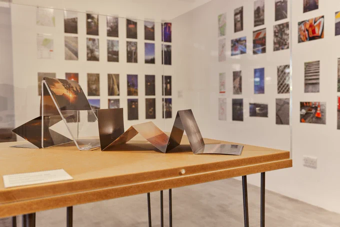Queering Connections: Triangulation exhibition at The Winchester Gallery. In the foreground a folded photobook is displayed on a wooden table. Photographs are displayed on white gallery walls in the background. Photo credit: Dave Gibbons