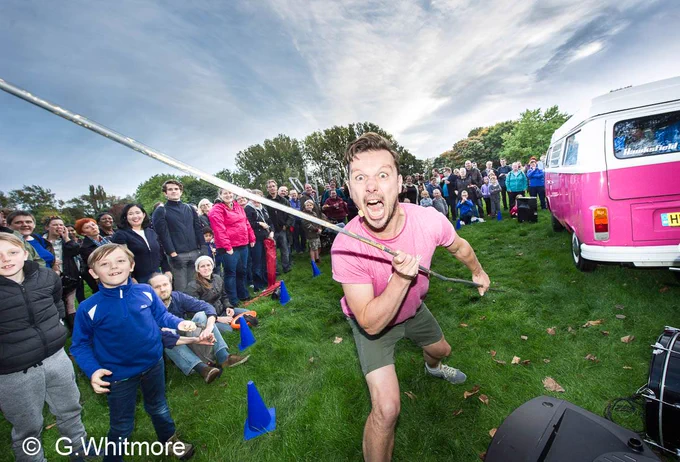 Tom, a white man with brown hair and a pink tshirt is shouting towards the camera with wide eyes and a wide mouth. He is yielding some sort of long stick which goes out of picture. Hi is outdoors, on grass and behind him is an audience of people of all ages smiling, and behind them are trees. To the right you can see the edge of a pink campervan.
