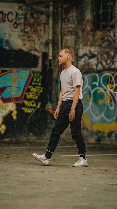 Connor is a young white male with short blond hair and beard. He is wearing a beige tshirt and black trousers and is standing in front of a graffitied wall.