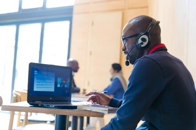 Picture of man wearing headphones and mic talking to tutor on a laptop in a communal space.
