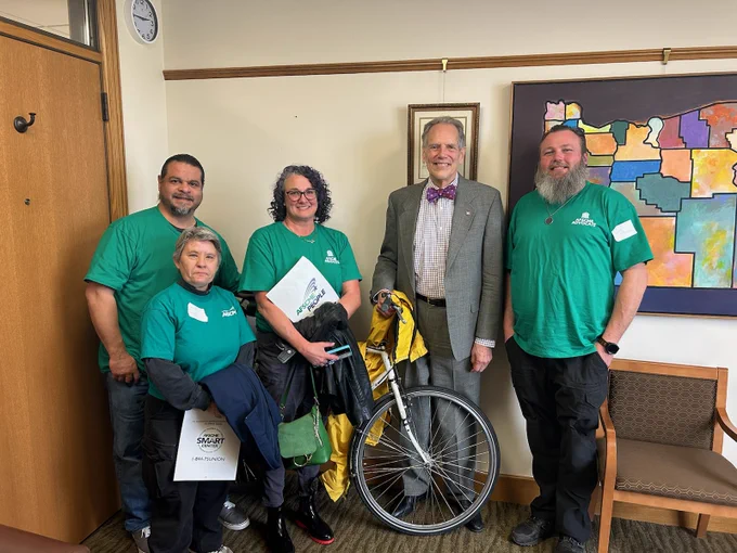 Representative Andersen wearing a gray suit with a purple bowtie in his office standing by his bike and four representatives from Oregon AFSCME wearing green shirts with the text "AFSCME advocate."