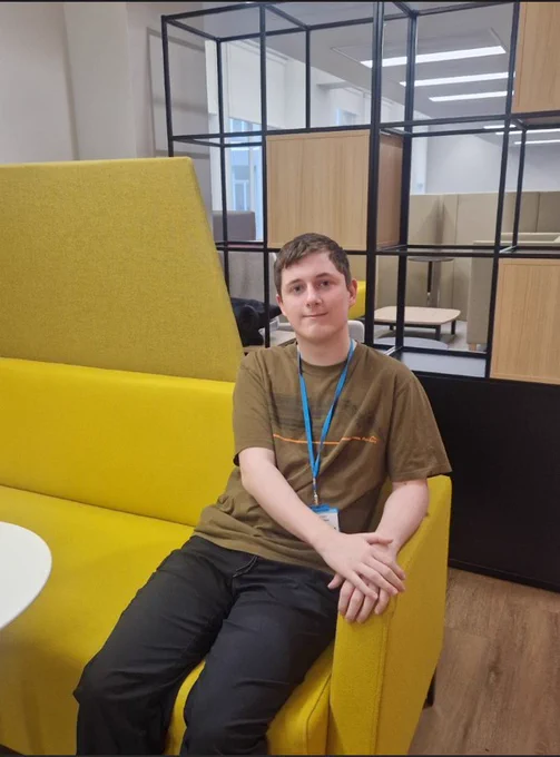 This is an image of an intern on our new HMRC supported internships. The image is of a young man wearing an olive t shirt, black jeans and a blue lanyard sitting on a yellow sofa, looking to camera. 
