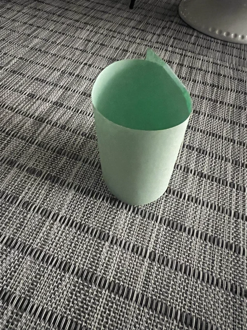 A paper “mug” made out of green paper by a 6 year old set on a gray placemat for  a “morning tea party”