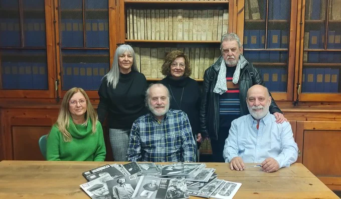 Fotografia de sis membres de l'Associació Plec a la sala de consulta de l'Arxiu Municipal de Lleida, situada als baixos del palau de la Paeria. Al fons es poden veure col·locats en una prestatgeria de fusta els exemplars dels llibres d'actes de La Paeria. A damunt de la taula es poden veure els exemplars de la revista Ple objecte de la donació. 
