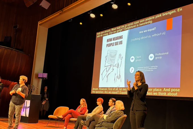 A group of people on a stage in front of a projected screen. Four of them are seated whilst two are standing and using BSL to communicate with the audience.