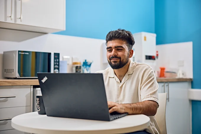 A smiling man sits at a kitchen table working on a laptop computer