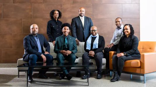 Seven Black men and women in business clothing (mostly suits), with three sitting on a couch, two on a chair, and two standing behind the couch