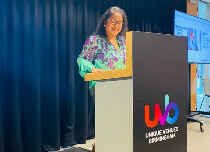A speaker stands at a Unique Venues Birmingham lectern, in front of a presentation screen and curtained backdrop. Wearing a green and purple top with black glasses and long black hair. 