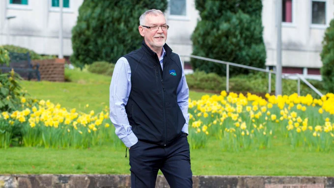 Ian Findlay CBE walks along a path, with a bed of daffodils on a verge and buildings behind him.