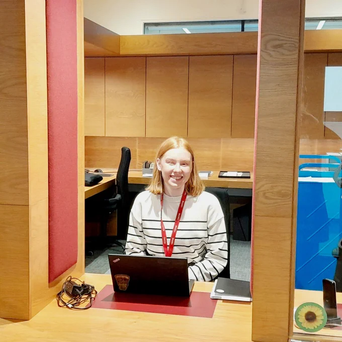 Photograph of Lauren Walmsley at the front desk of the modern reading room at John Rylands Library. Lauren is sat behind a laptop and is smiling at the photographer.
