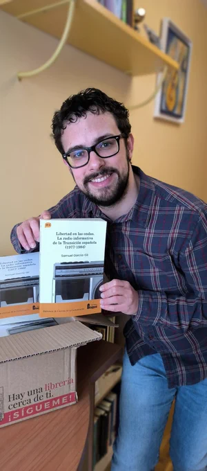 El autor posando orgulloso con una caja llena de ejemplares del libro, mientras sostiene uno de ellos a cámara.