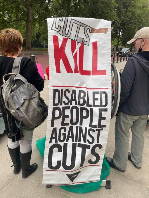 Activists stand with a banner between them image devious a cleaver that spells out cuts with kill in red then disabled people against cuts underneath in black