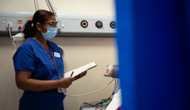 Photo of a nurse wearing a face mask and talking to a patient who is hidden behind a blue hospital curtain. The nurse is holding a clipboard and a pen in her hand and standing next to a monitoring machine.