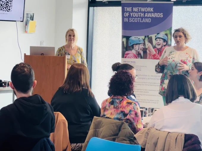 Kate Still stands at a podium delivering a presentation. To the right is a pop-up banner for the Awards Network.