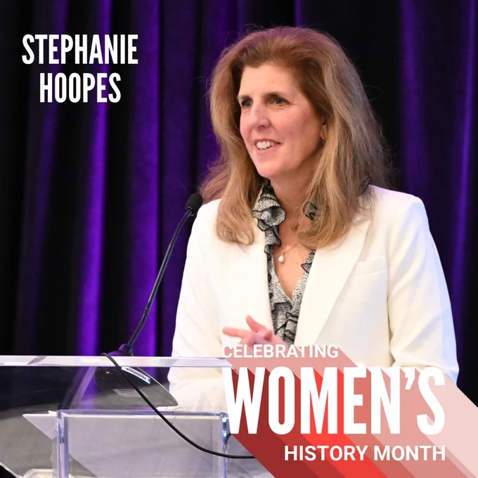 Stephanie Hoopes stands at a glass podium, wearing a white blazer and smiling at an audience. Text reads: Celebrating Women's History Month, Stephanie Hoopes