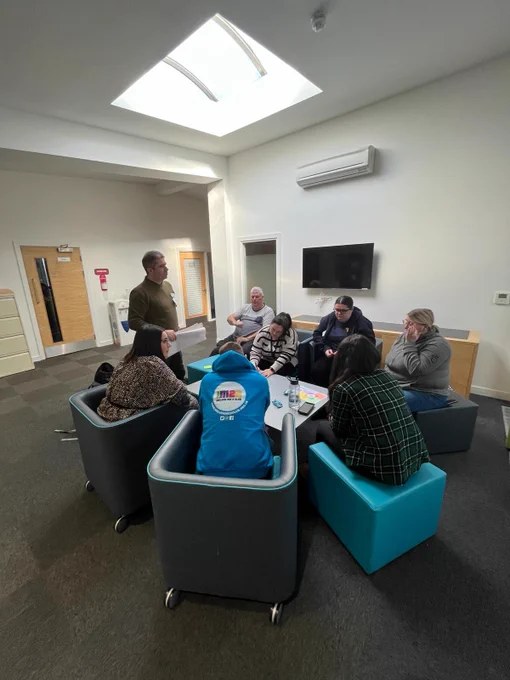 A group of attendees sit in a circle around a table. Above them is a large skylight.