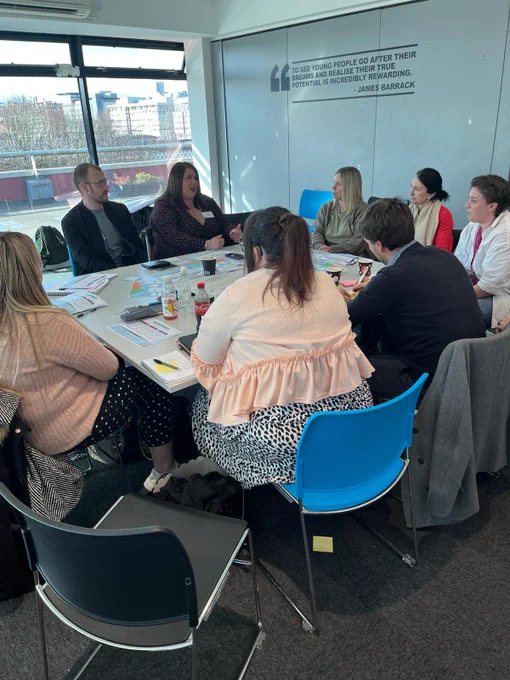 A group of attendees sit around a large square table. They are engaged in discussion.