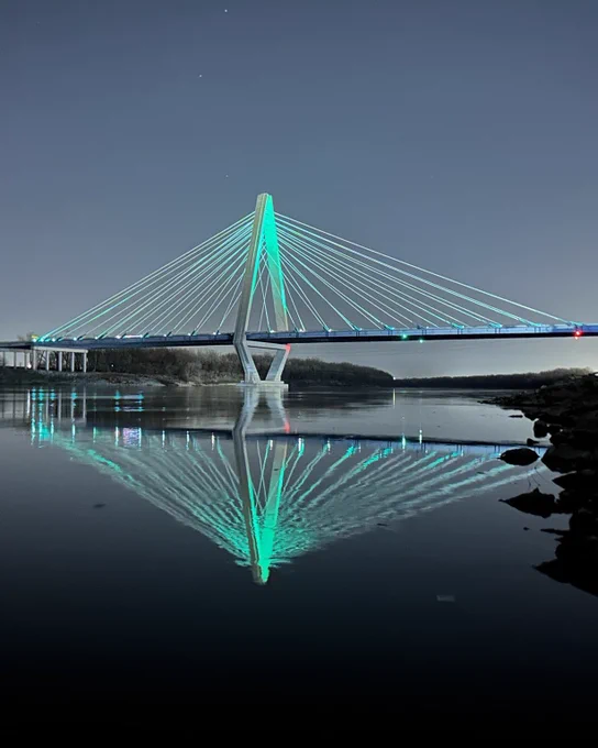 Photo of Bond Bridge lit up teal with its reflection showing on the Missouri River