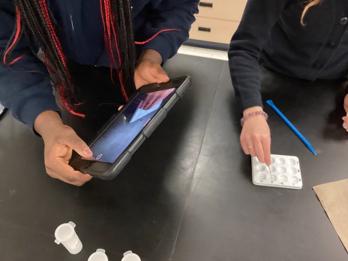A student using a toothpick to stir a mixture in a spotwell while another student records the results using an iPad mini.