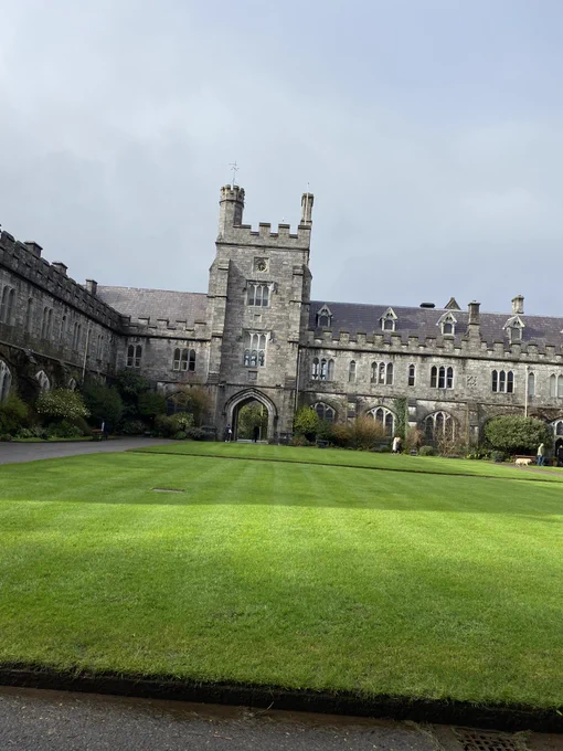 Image of UCC's Main Quadrangle, a castle style building with a walk-through archway to the left of the north-wing of the building. Light grey and white clouds line the sky, as the vibrant green grass of the quad set the foreground of the image. 