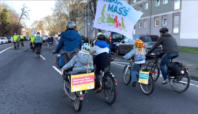 KidicalMass Kinder, Eltern, Familien auf Fahrrädern mit Demoschildern "Macht uns Platz, sonst gibt's Rabatz" und KidicalMass Fahne auf mehrspuriger Stadtstraße
