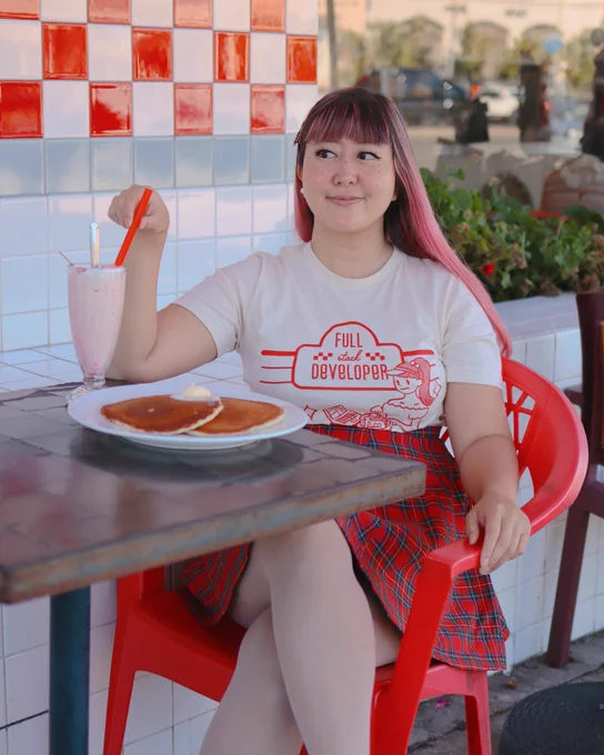 me, a feminine person with pink hair, wearing a shirt that says “full stack developer”, while sitting at an outside table of a diner, eating pancakes and drinking a milkshake
