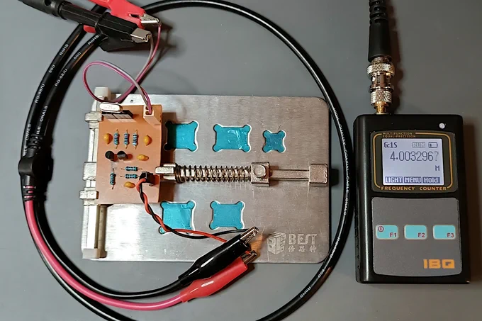 The home-made Oscillator circuit held on a spring-loaded PCB vice with power connected and the output being monitored by a small handheld radio frequency counter. A Crystal is inserted in the black single row socket at the top, and the frequency counter screen reads 4.0033 Mhz