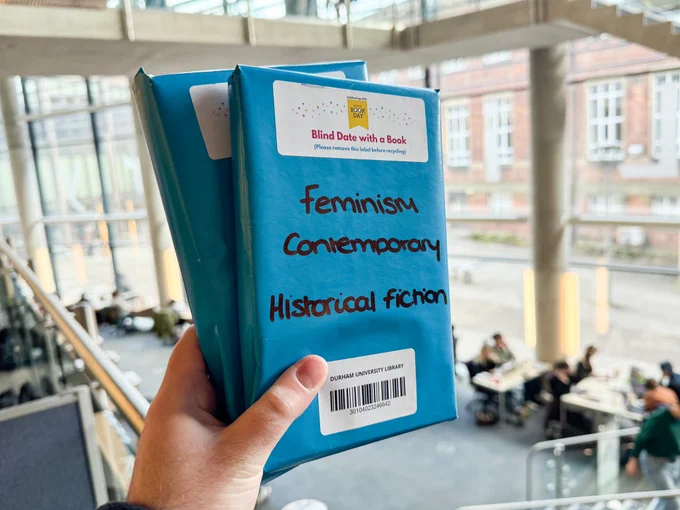 A hand holding up two World Book Day books wrapped in blue paper. The Library East Wing atrium is in the background.