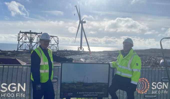 Two people wearing protective clothing standing in front of a construction site with a turbine in the background.