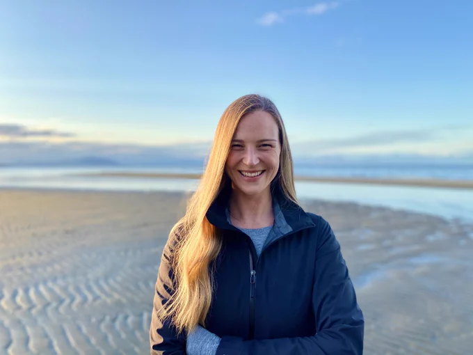 Alix Shield with long blonde hair, wearing a grey sweater and blue jacket, stands smiling on a sandy beach in front of a body of water. 