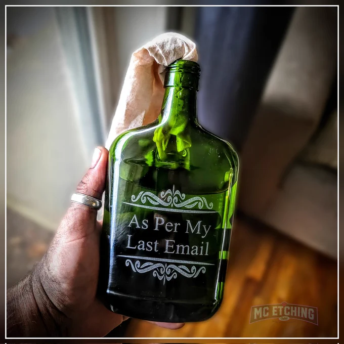 Hand holding a green glass bottle with a cloth sticking out of the mouth. Upon the bottle is etched the phrase "As Per My Last Email." With a top and bottom filigree accent border. 