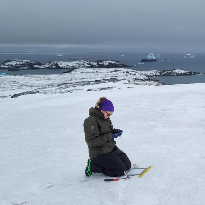Emily kneels in the snow taking samples. The landscape falls away behind her - into rocky outcrops, and then cold Antarctic sea with icebergs.