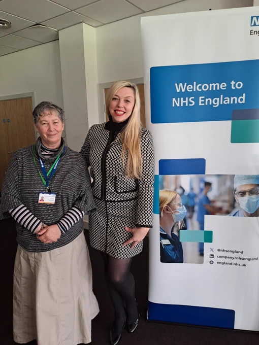 Stacey and Tina, smiling at the camera and standing next to a 'Welcome to NHS England' banner.