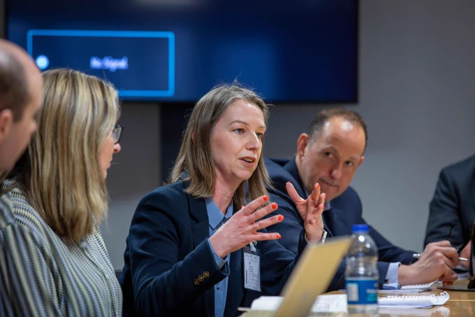 Chair of The Pension Protection Fund, Kate Jones is seated at the roundtable, speaking and gesturing with her hands in front of her. Two other attendees are visible in the foreground and background, to the left and right.