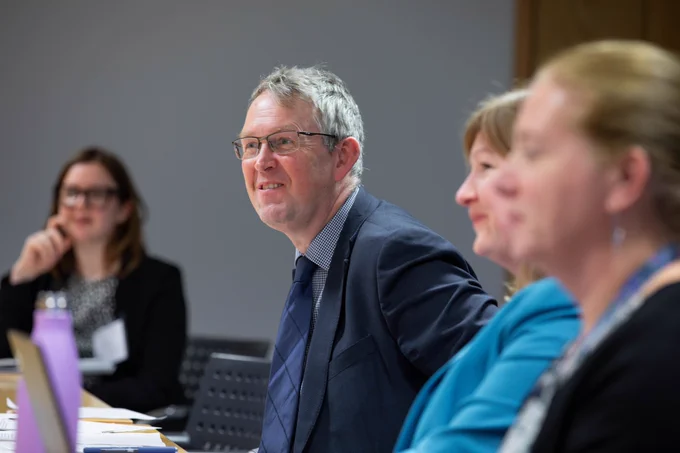 The Minister for Pensions is also seated at a table. He is looking across the table, intently listening to someone speaking who is out of view. Other attendees are sitting to his left and right, also listening to the speaker.