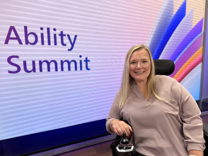 Jessica in her power wheelchair smiles in front of a large screen that says Ability Summit