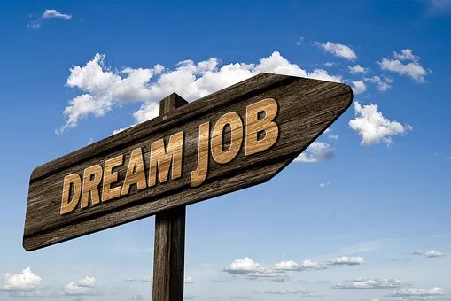 Image of a wooden sign with the words 'dream job' on it. In the background there is blue sky with white fluffy clouds.
