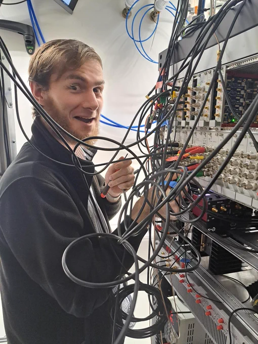 A man is standing in front of an electronics rack holding a screwdriver and looking as though he's about to make some changes to a board. There are lots of wires heading in all directions.