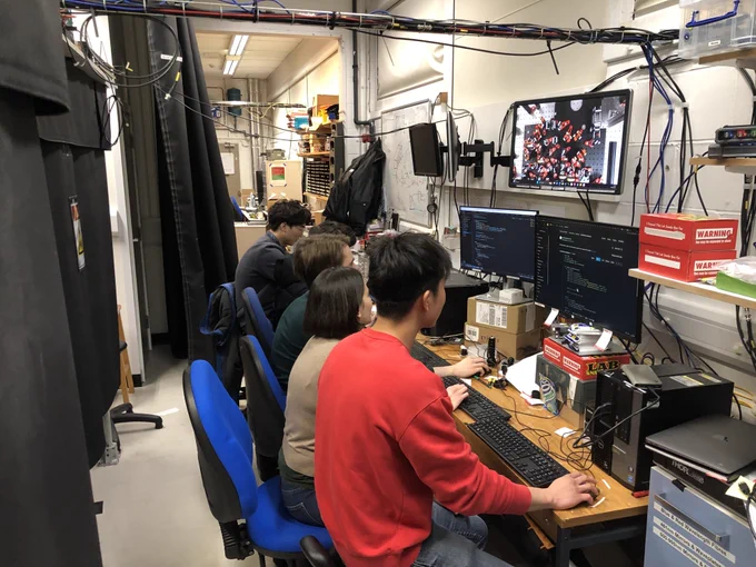 Some people are sitting at a long wooden desk in a laboratory, all looking intently at computer screens showing the code they're writing to control the experiment.