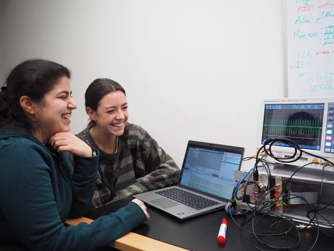 Two women are sitting at a desk looking at an oscilliscope screen which is displaying a sine wave. A laptop is open in front of them and the oscilloscope is connected up to a crate with various wires and lights. They look very happy with their results!